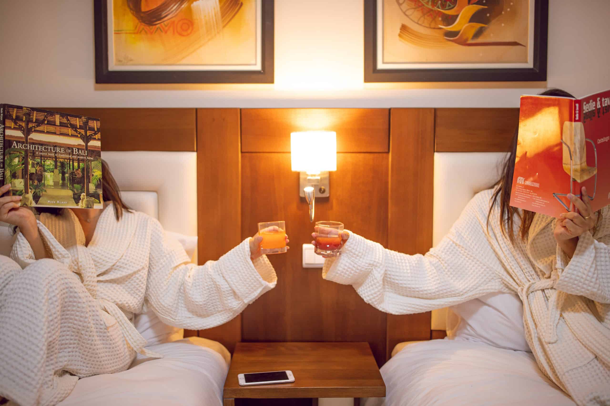two friends sit on a twin bed each and make a toast while reading a magazine. That magazine is covering their faces.